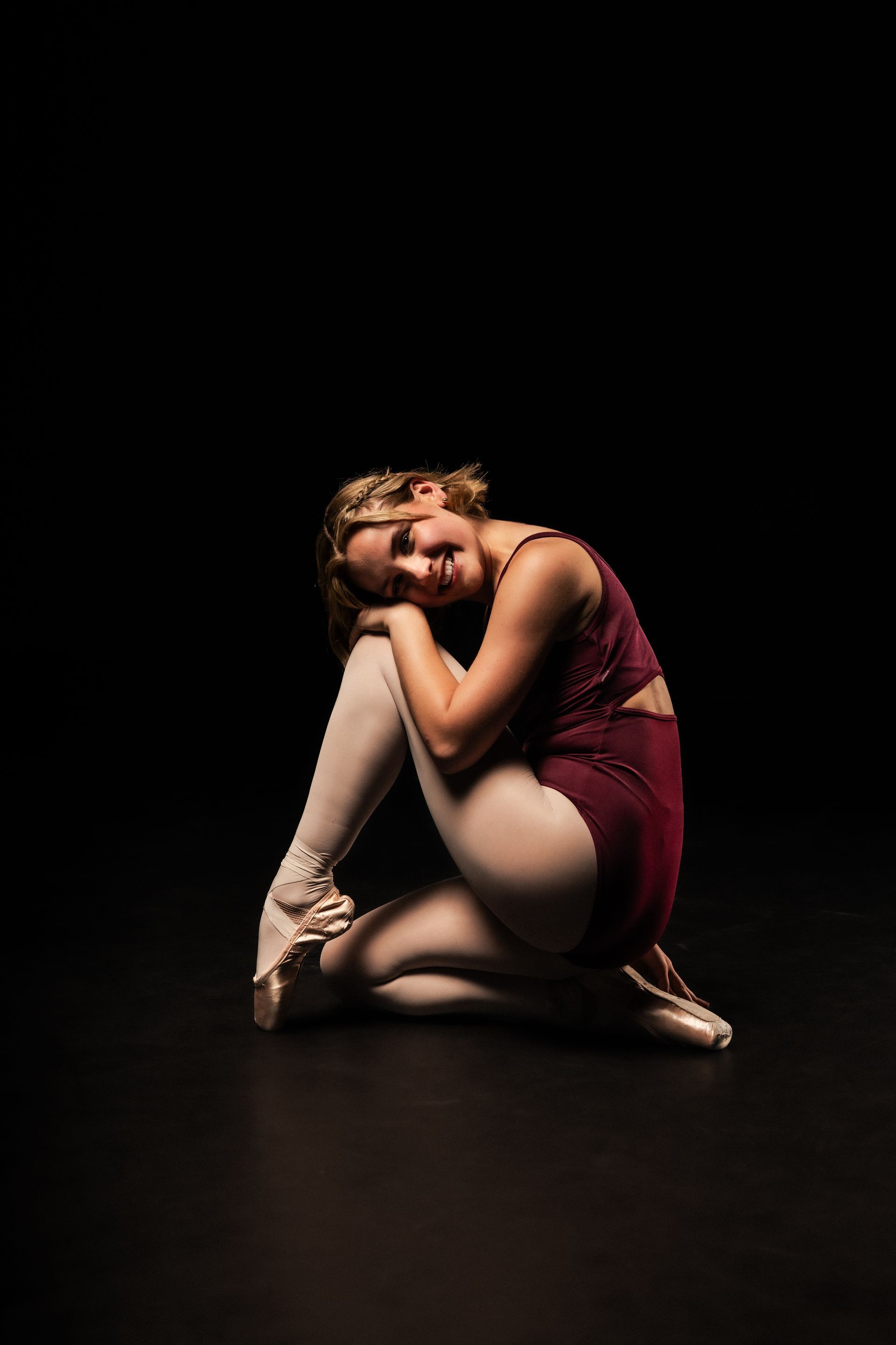 Dancer in a burgundy costume crouches on a dark stage, resting her head on one hand.