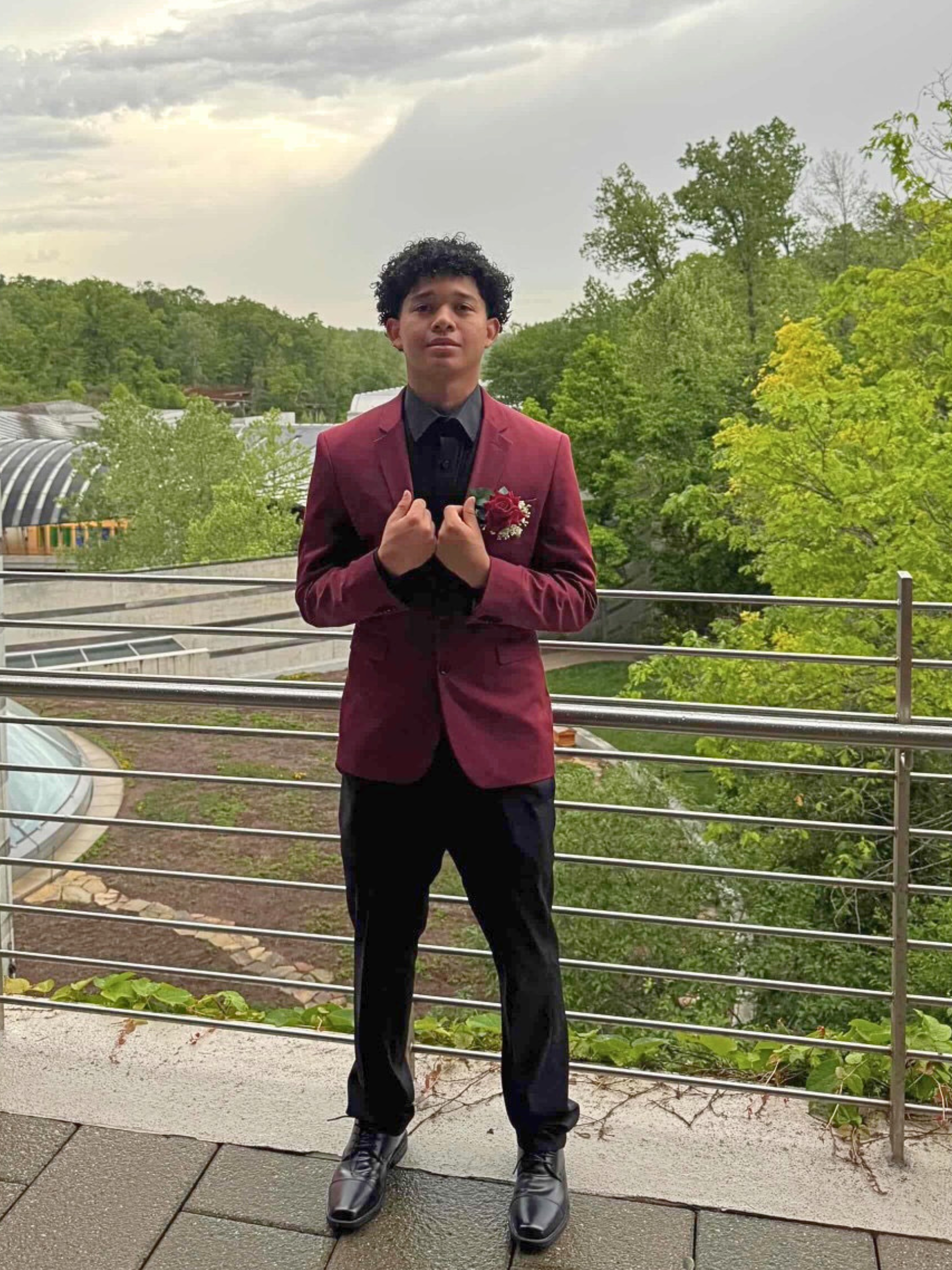 Person in a maroon blazer standing by a metal railing in a lush park, holding their hands near their chest.