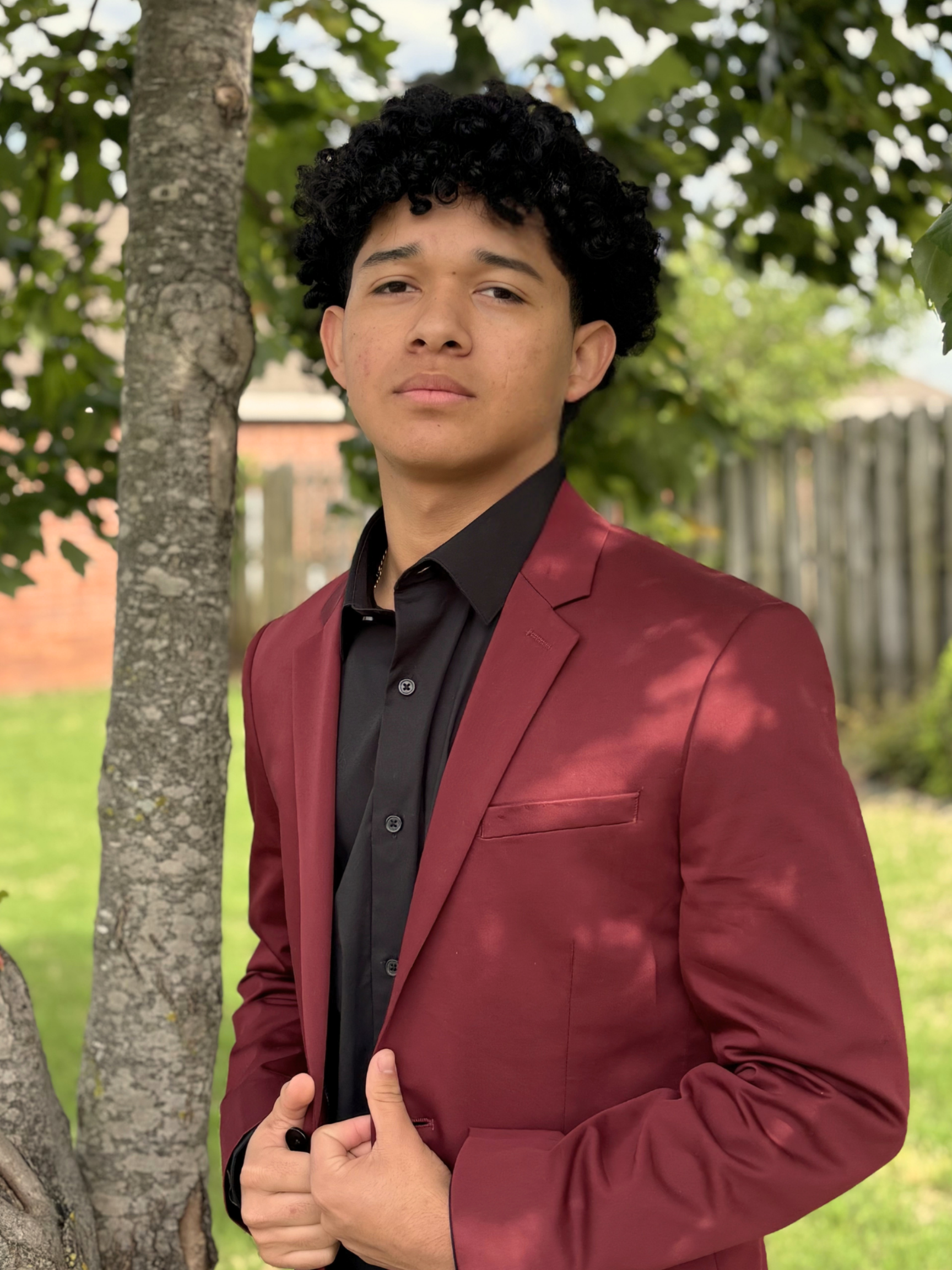 Person in a burgundy blazer standing by a tree outdoors, looking at the camera.
