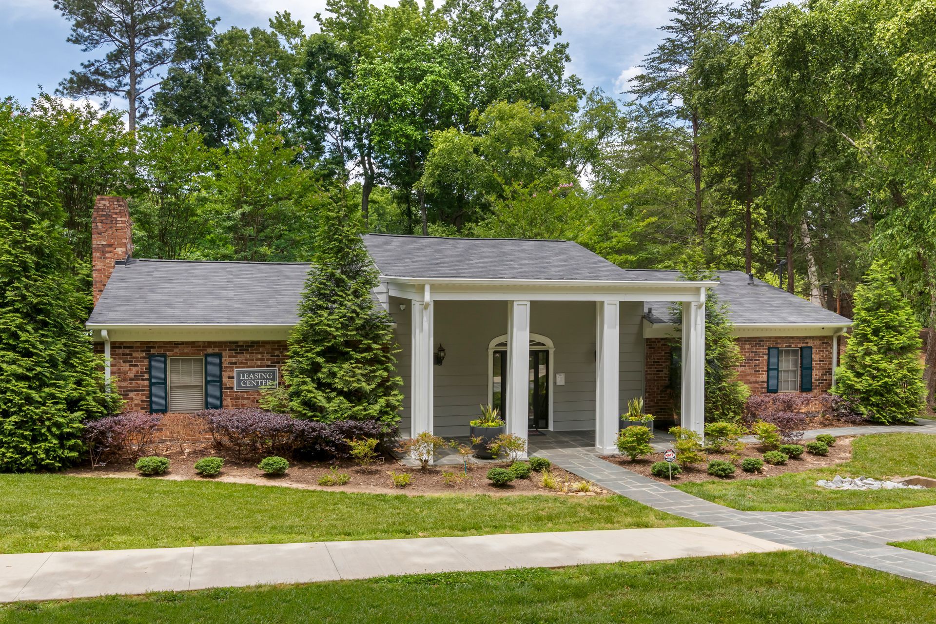 Ranch-style house with brick and gray exterior, pillars, surrounded by greenery and lawn.