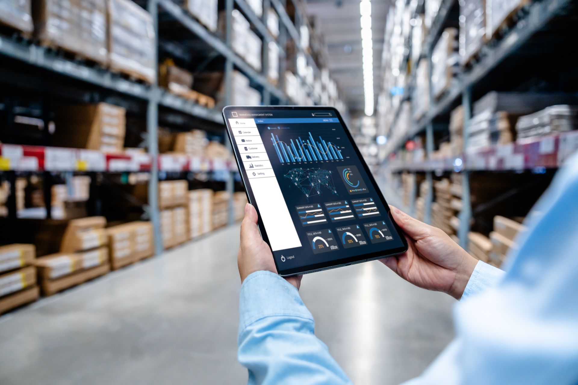 Person holding a tablet with data displayed, in a warehouse aisle with packed shelves.