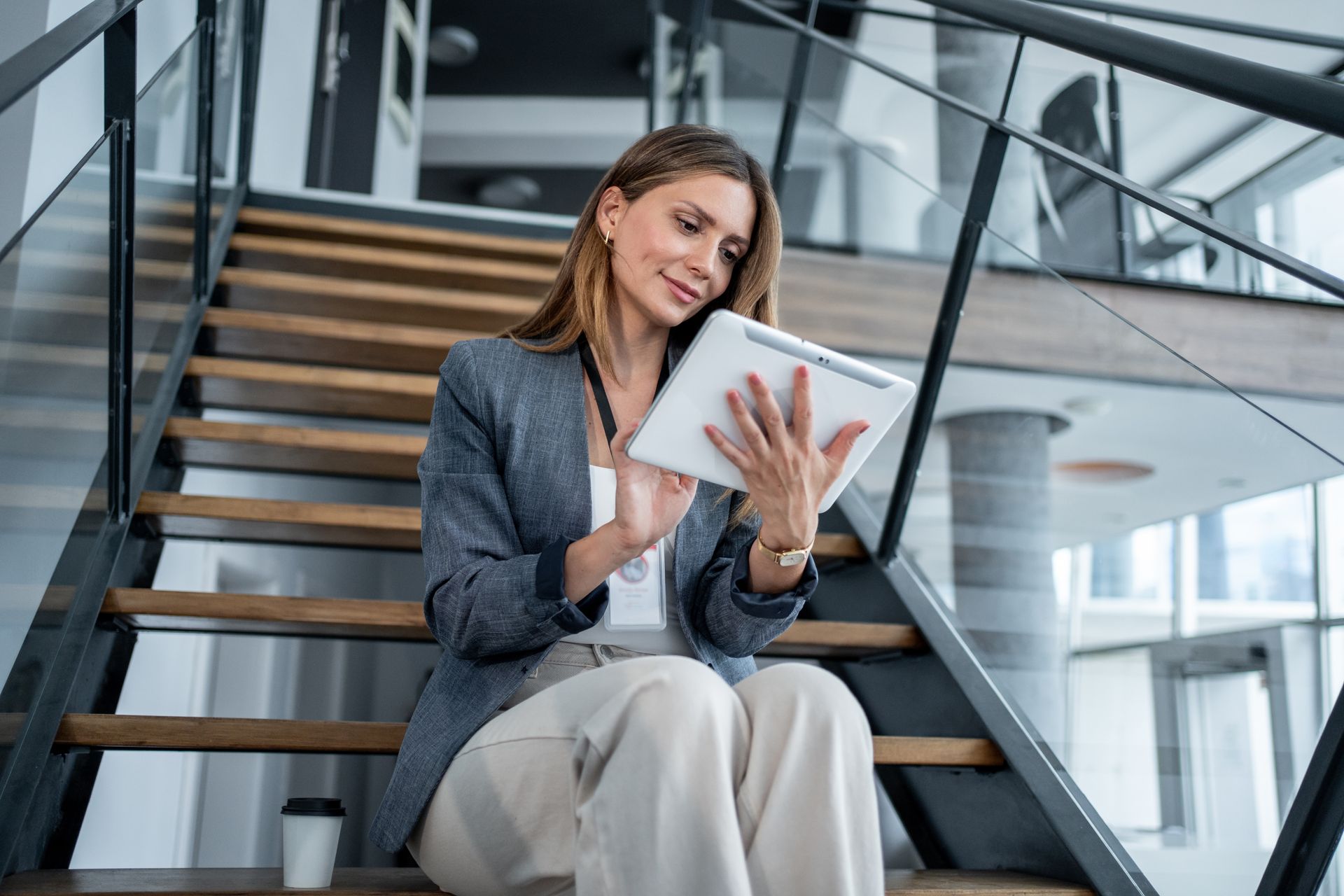 Woman in a gray blazer sits on wooden stairs, looking at a tablet. A coffee cup sits next to her.