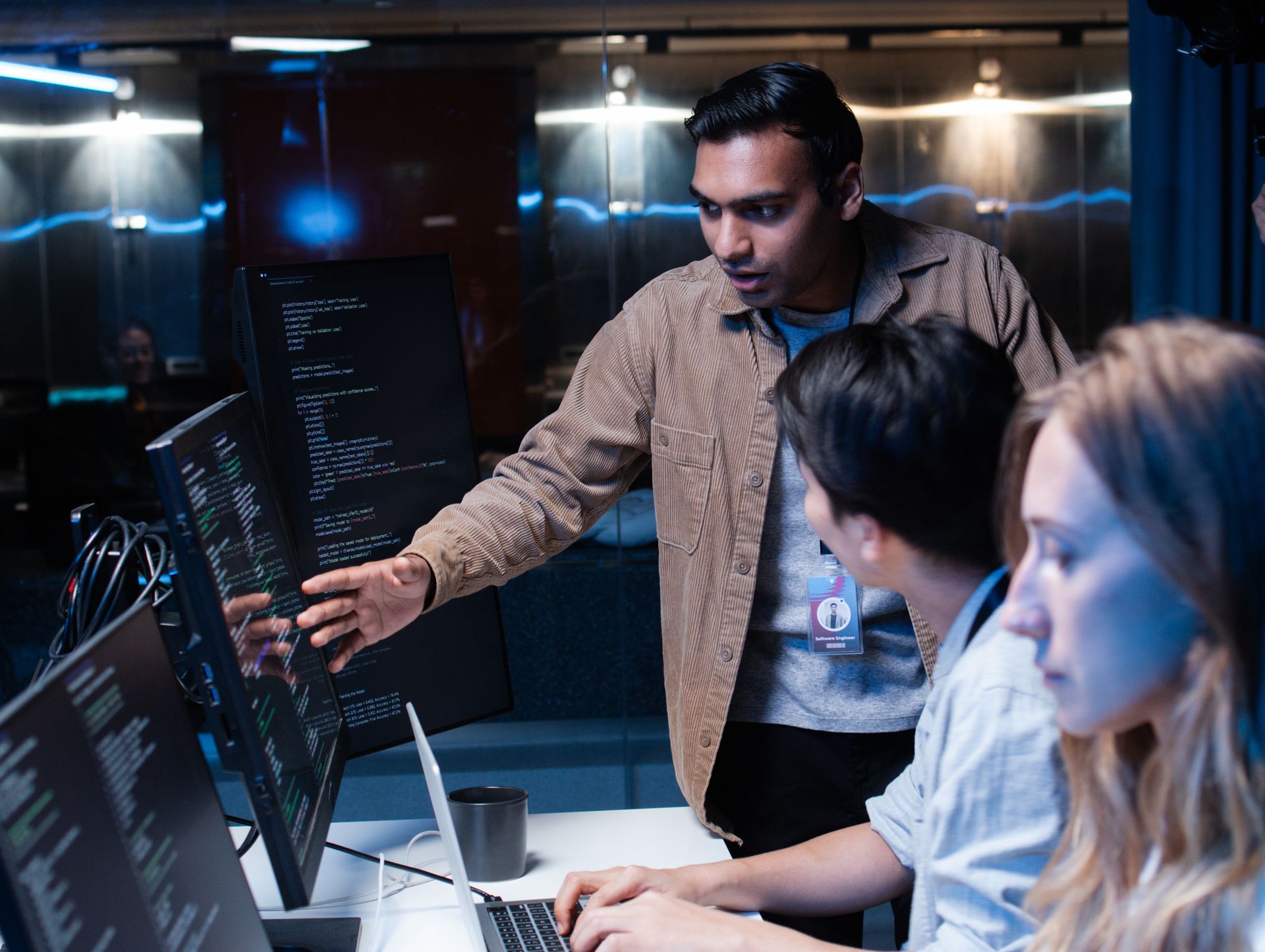Man pointing at computer screen, explaining code to two colleagues in a dark, tech-filled office.