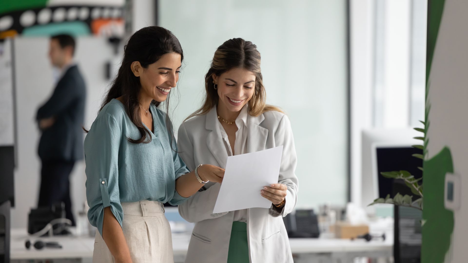 Two women in an office looking at a document and smiling, another person is blurred in the background.