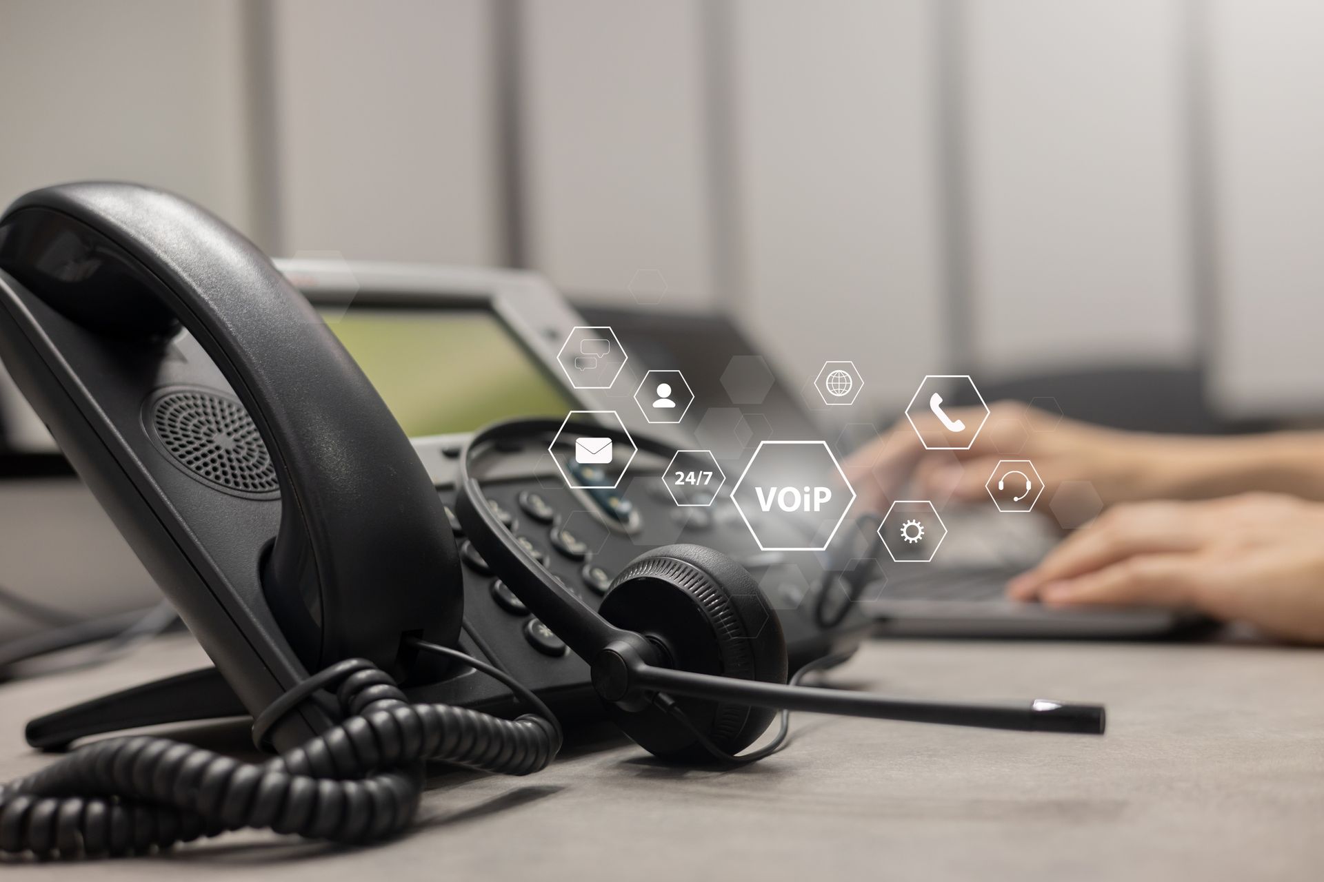 Black office phone with headset and VoIP graphic overlay. Hands on laptop in background.