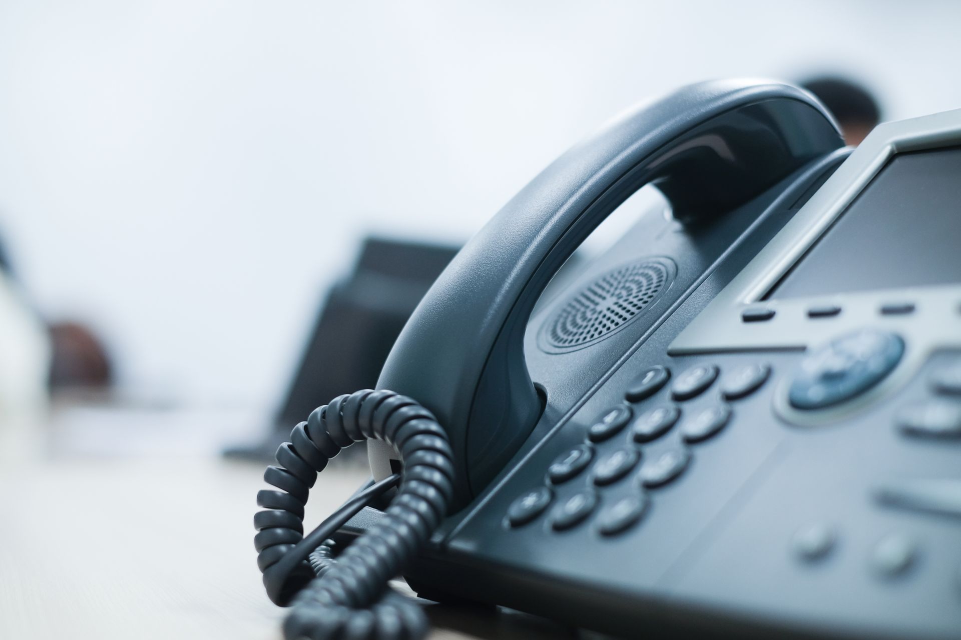 Close-up of a dark gray office telephone, handset up, on a desk with a blurred background.