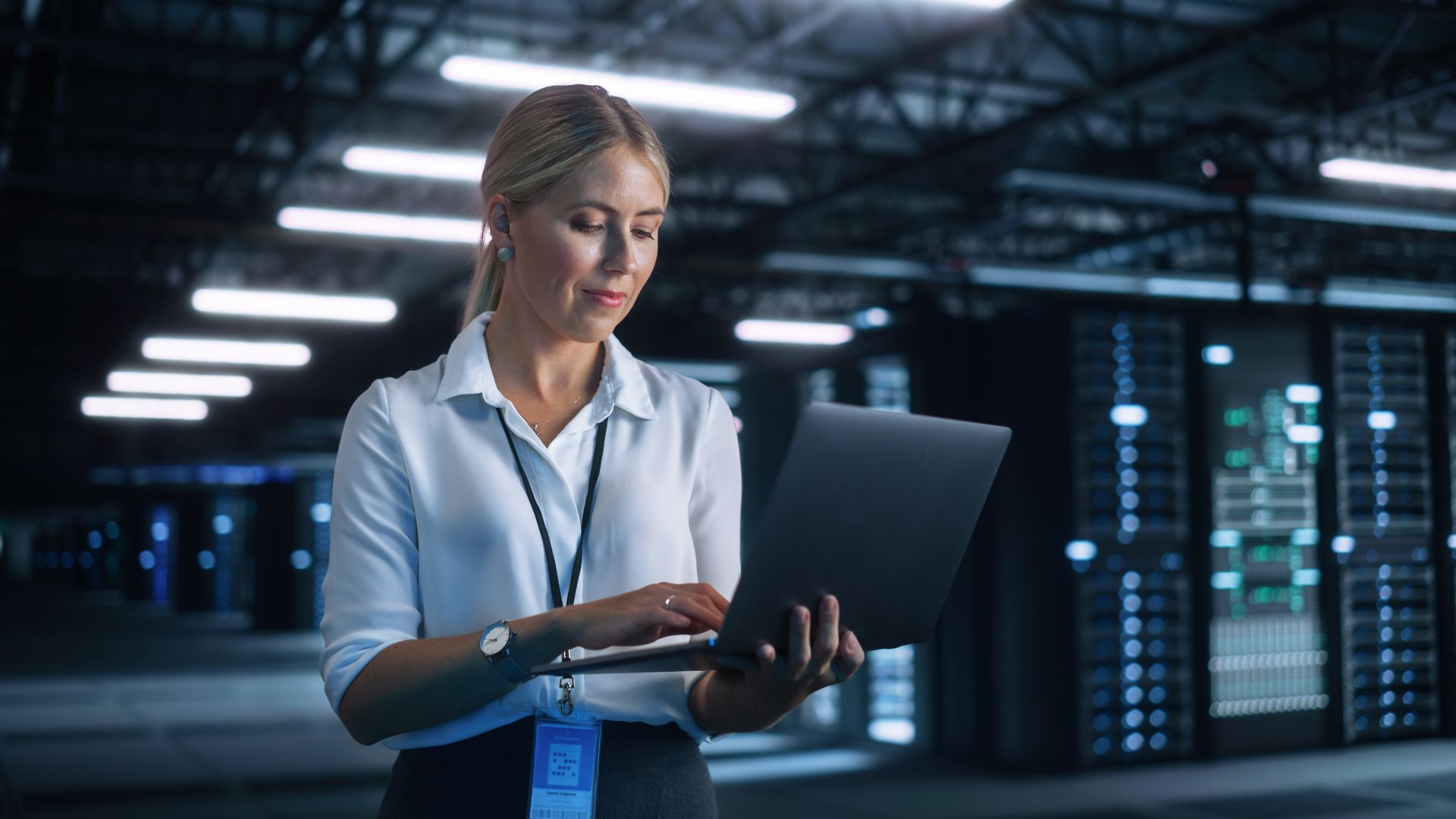 Woman working on laptop in a server room with racks of computers.