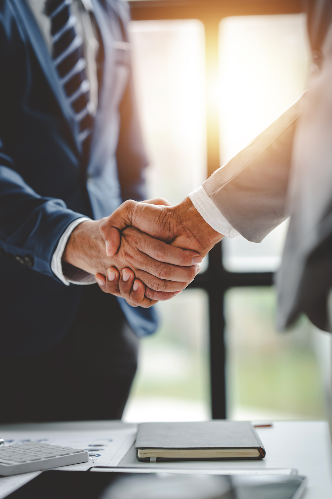Two people in suits shaking hands at a desk, business meeting.