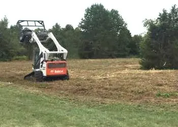 A bobcat is cutting grass in a field with trees in the background.