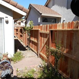 Narrow side yard with a red wooden fence, gravel path, and small shrubs between houses