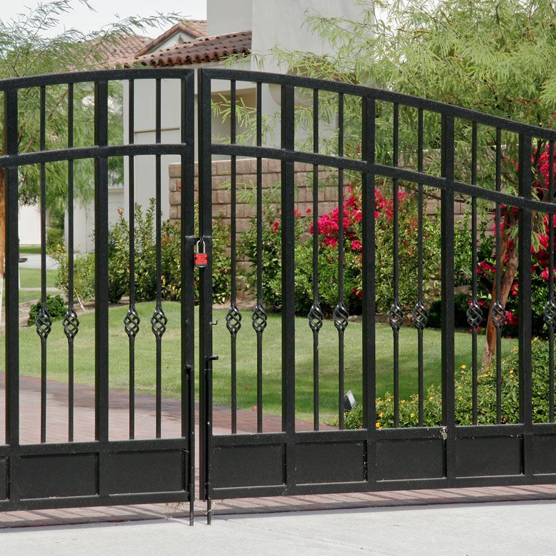 Black wrought iron driveway gate with decorative bars in front of a garden and house.