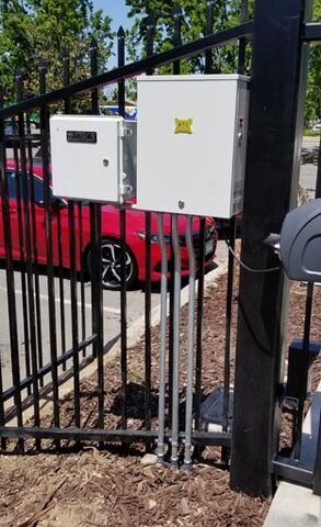 Two white utility boxes mounted on a black wrought iron fence, with a red car parked behind them.