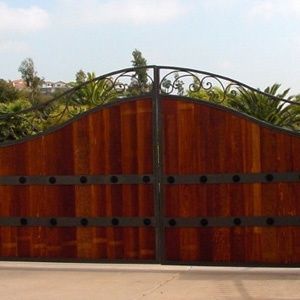 Large wooden gate with black wrought-iron top details, set against a garden and sky.
