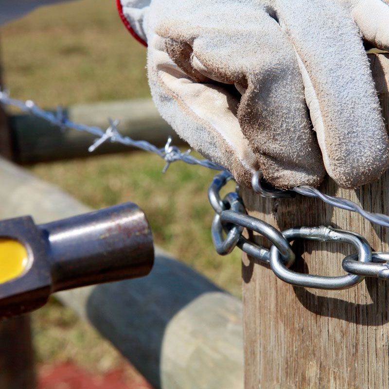Gloved hand fastening barbed wire to a wooden fence post with a chain and tool