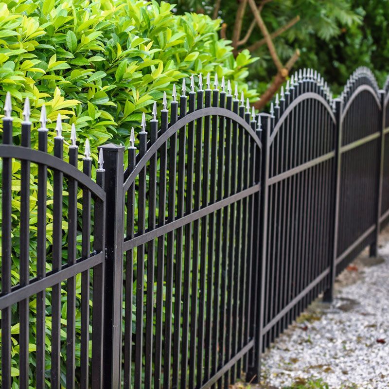 Black arched wrought iron fence beside green hedges and a gravel path