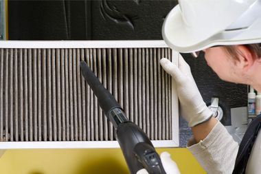 A man wearing a hard hat and gloves is cleaning a filter with a vacuum cleaner.