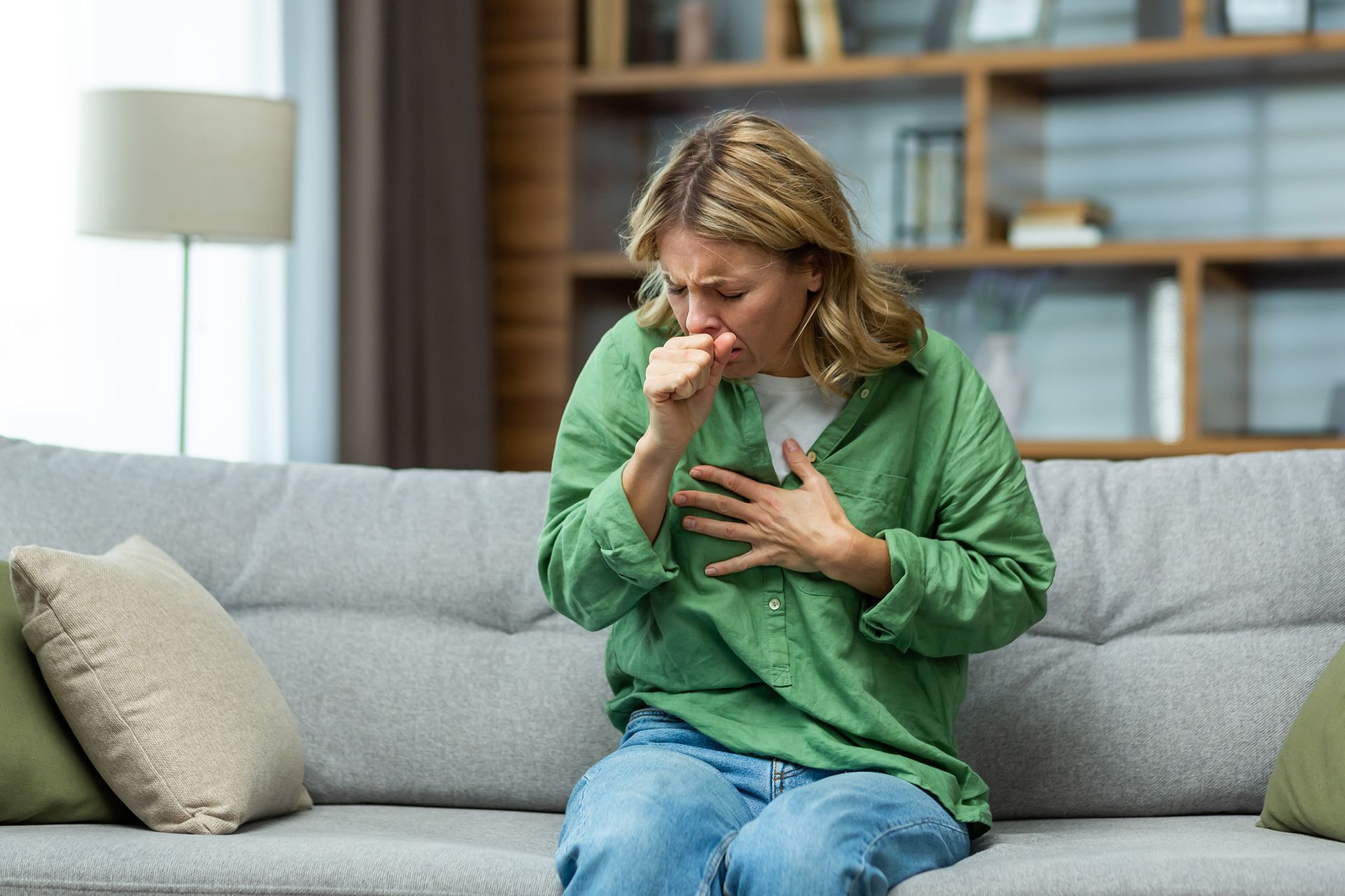 A woman is sitting on a couch coughing and holding her chest.