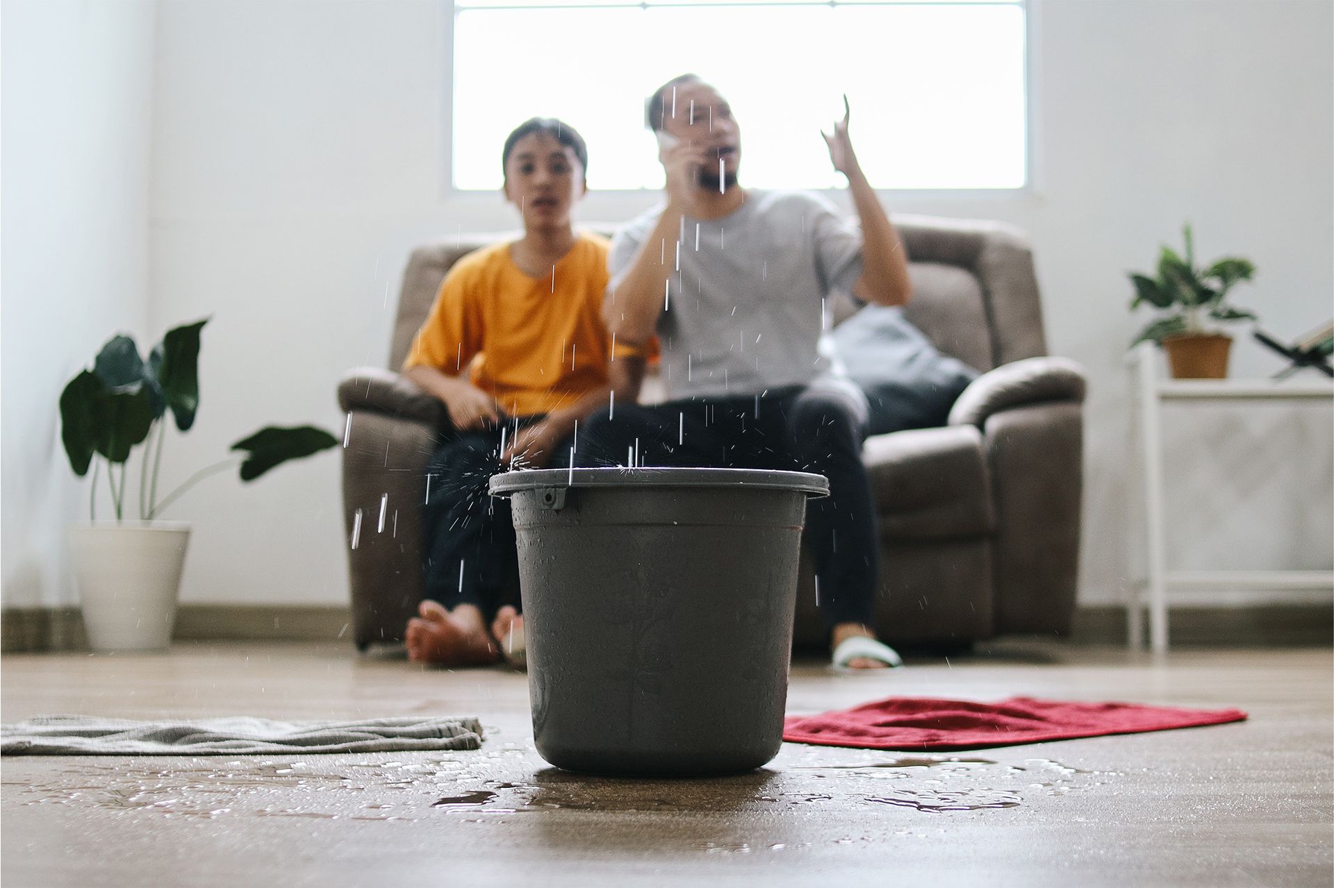 A man and a boy are sitting on a couch in a living room with a bucket of water on the floor.