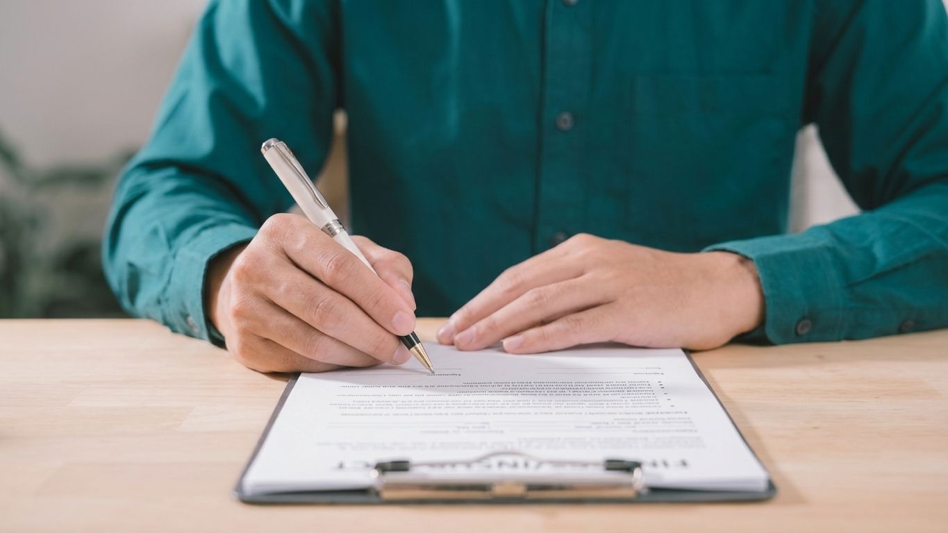 Hand writing on forms beside a blue umbrella, house model, car, and coins, suggesting home insurance