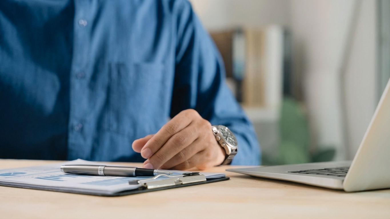 Hand placing a white umbrella over a model house on a desk beside a laptop