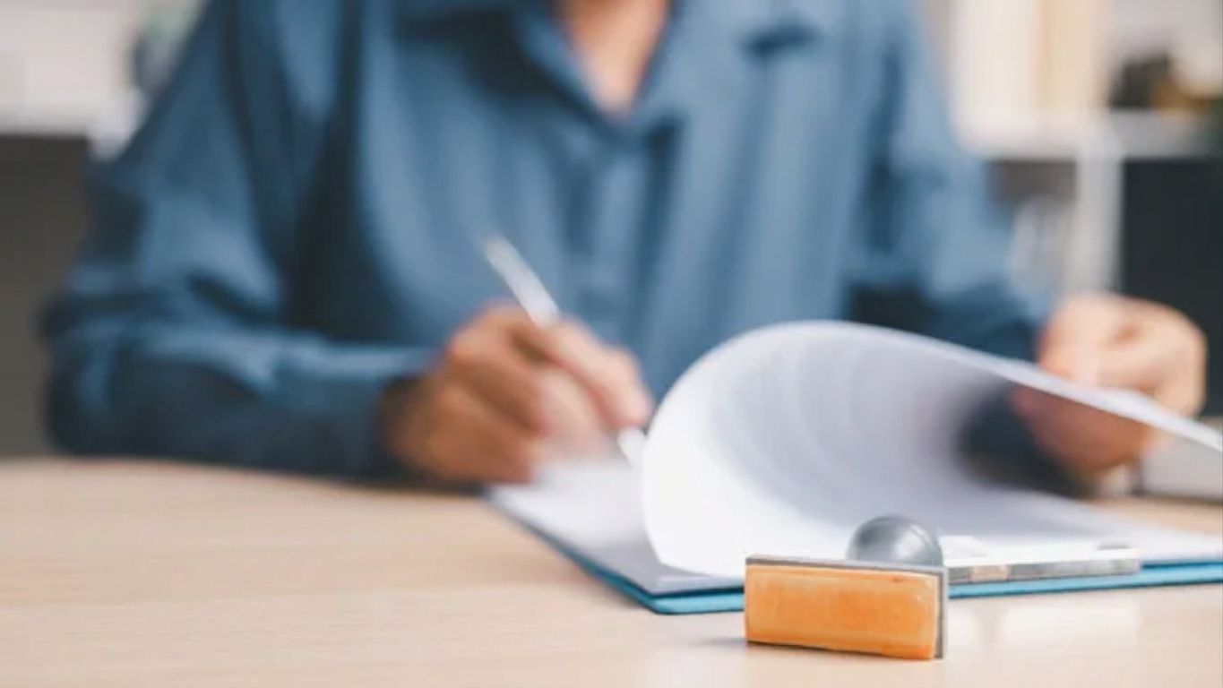 Small house model and key on a desk with a person signing paperwork in the background