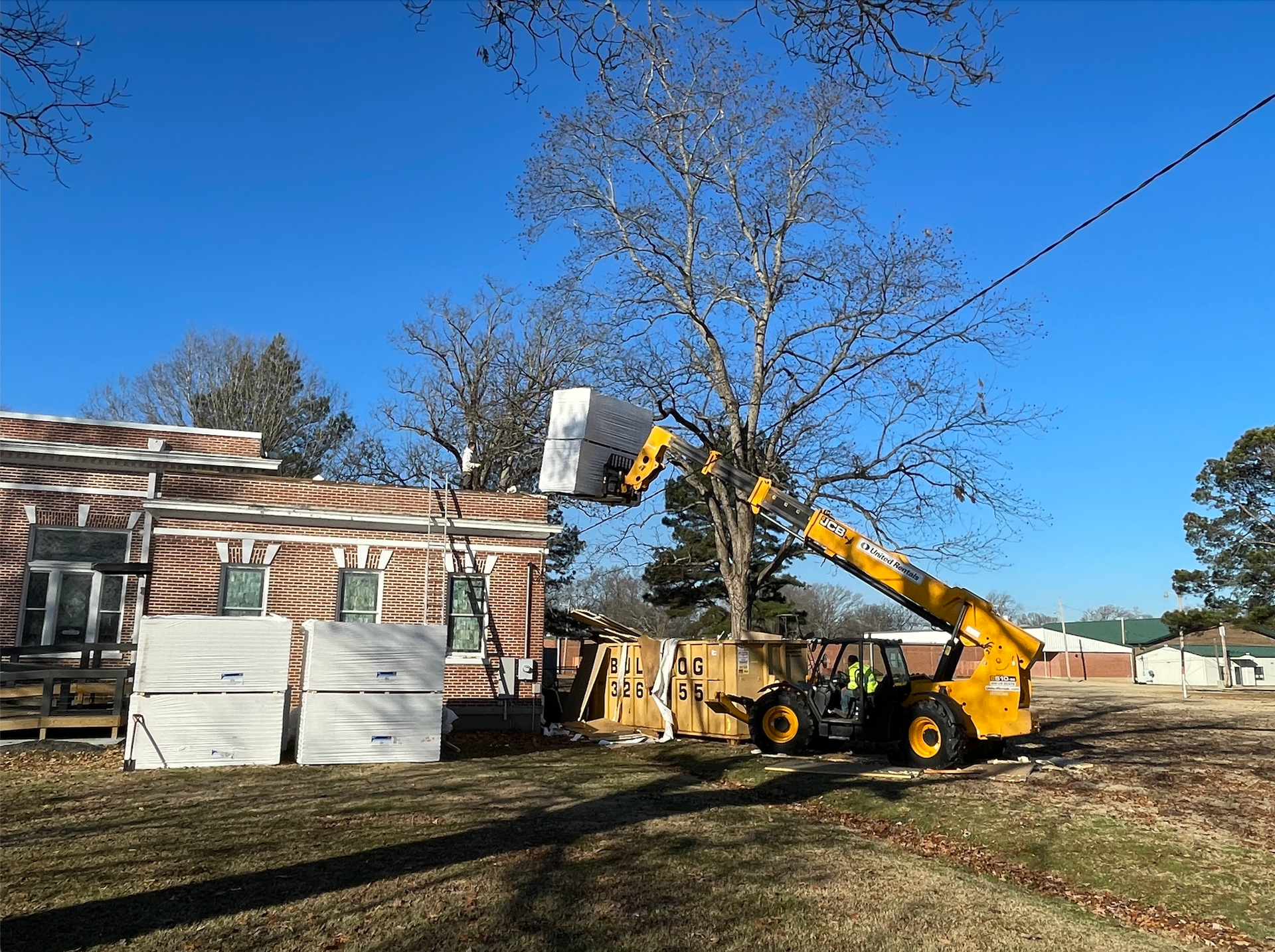 Yellow excavator lifting debris beside a log cabin under a clear blue sky