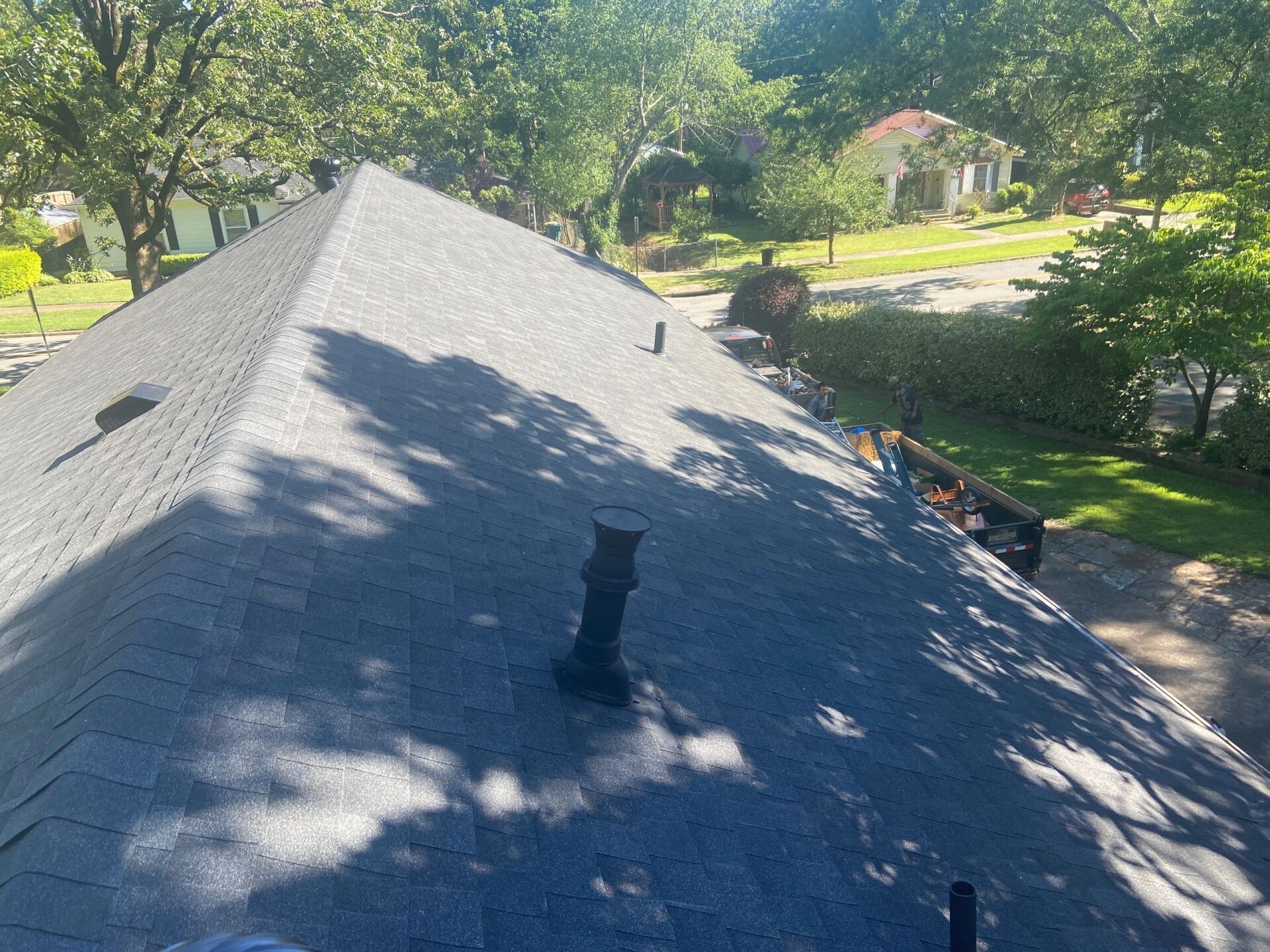 Gray shingled roof with a chimney, viewed from above, surrounded by trees and a sunny yard