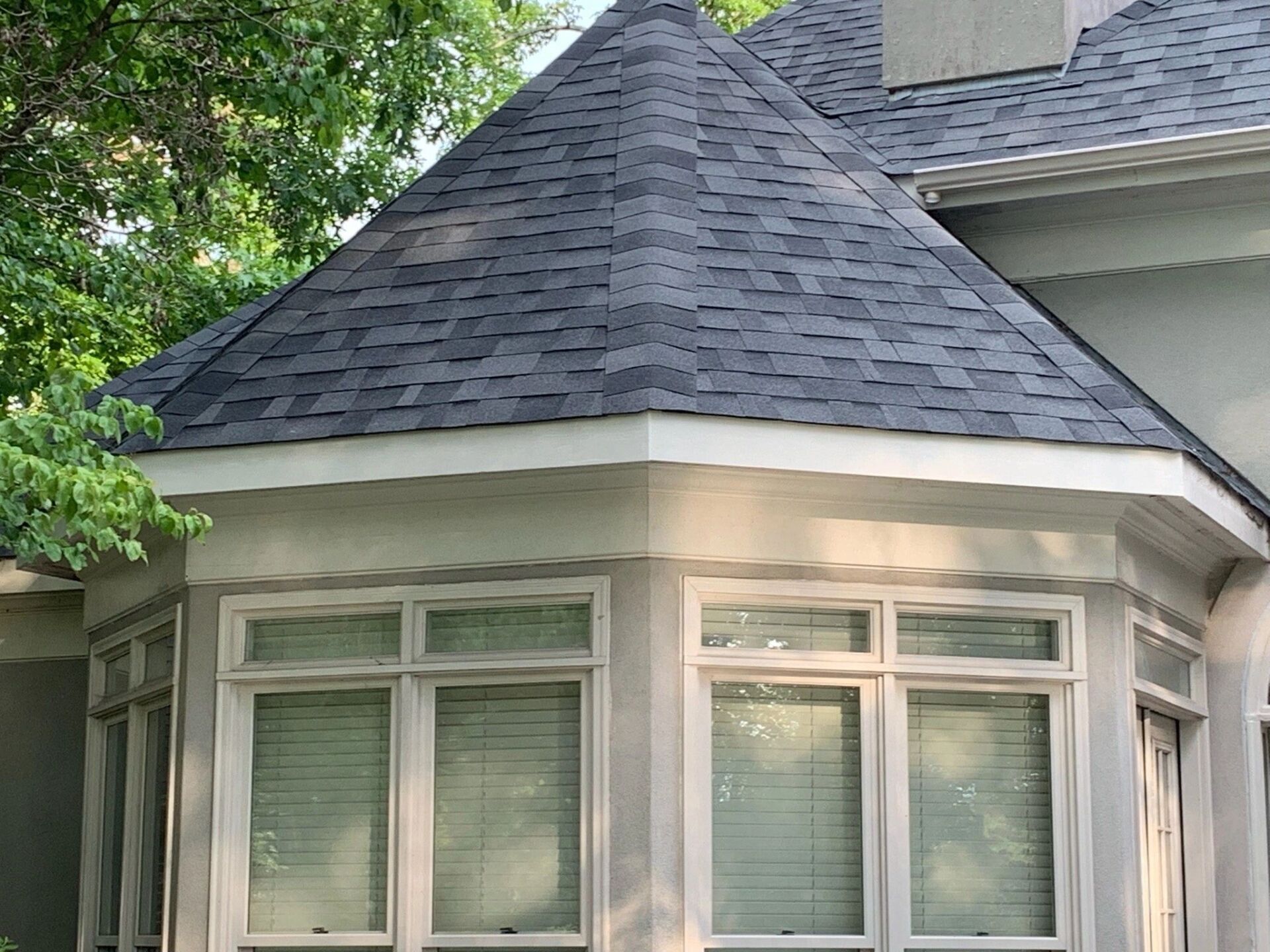White bay window with dark shingled roof and surrounding trees on a house exterior