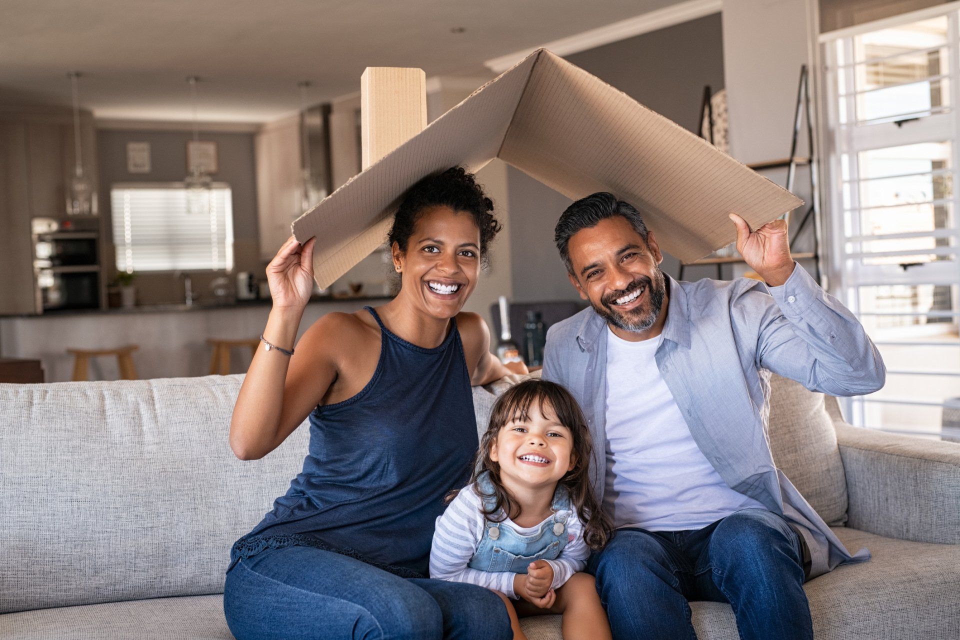 Family sitting on a couch under a cardboard roof, smiling in a bright living room.