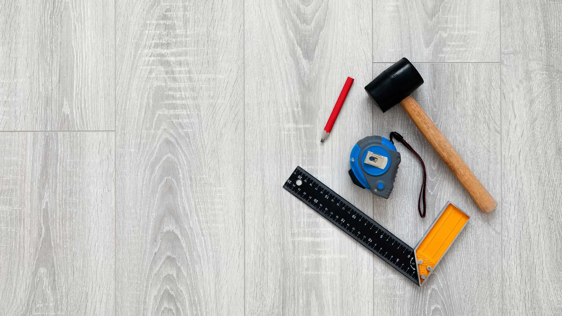 A hammer , tape measure , ruler , and pencil are on a wooden floor.