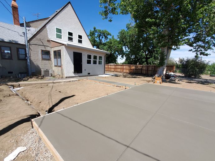 Fresh concrete patio beside a house under construction, with dirt yard and a large tree nearby