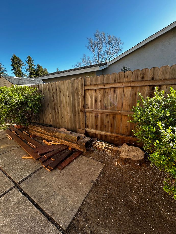 Backyard fence with stacked wooden boards, concrete patio, and shrubs under a blue sky