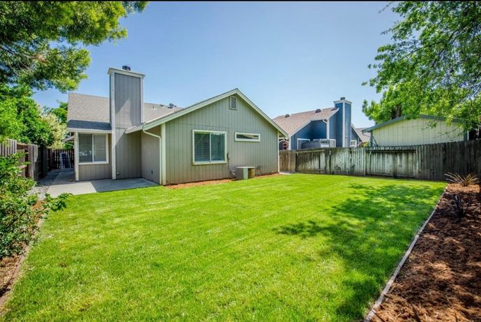 Backyard with green lawn, single-story house, wooden fence, and trees on a sunny day