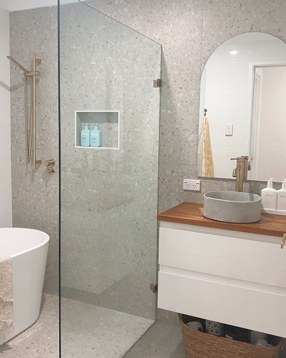 Modern bathroom with gray speckled tile, glass shower, gold fixtures, and wooden vanity.