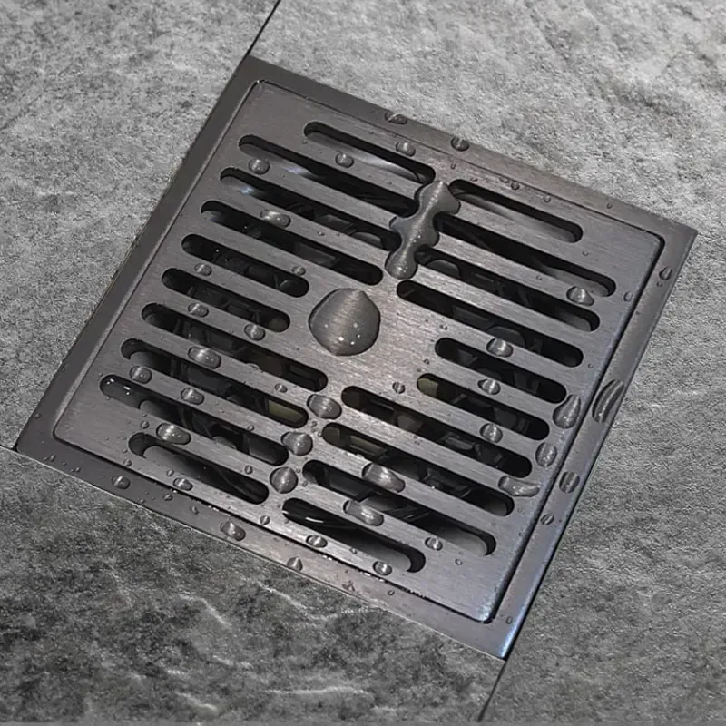 Square stainless steel shower drain with water droplets on gray tiled floor.