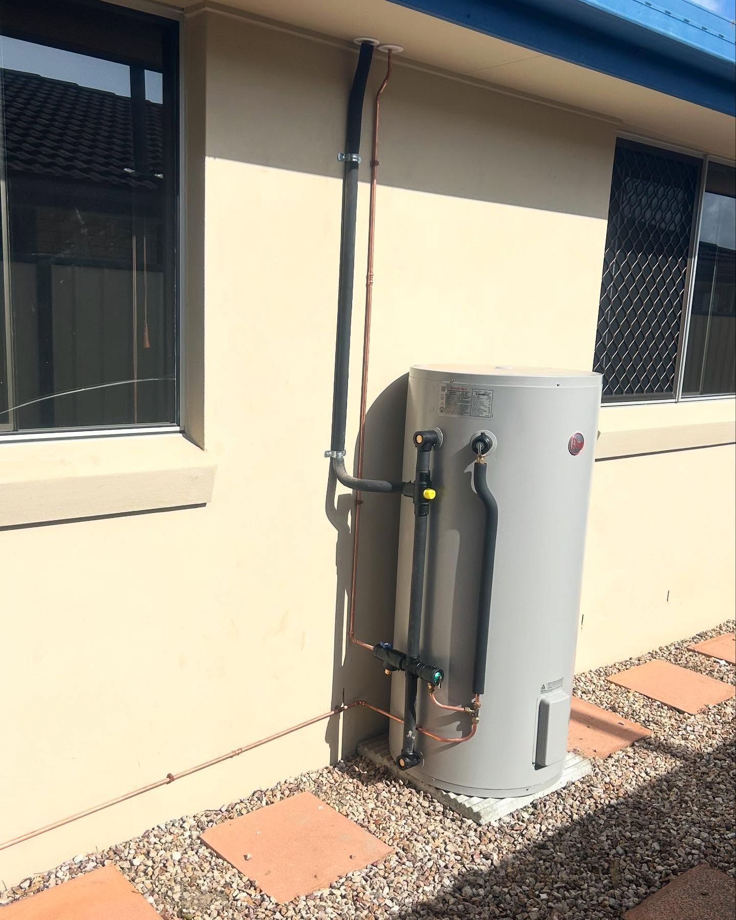A grey water heater against a beige wall, with black pipes. It sits on a concrete pad with surrounding gravel.