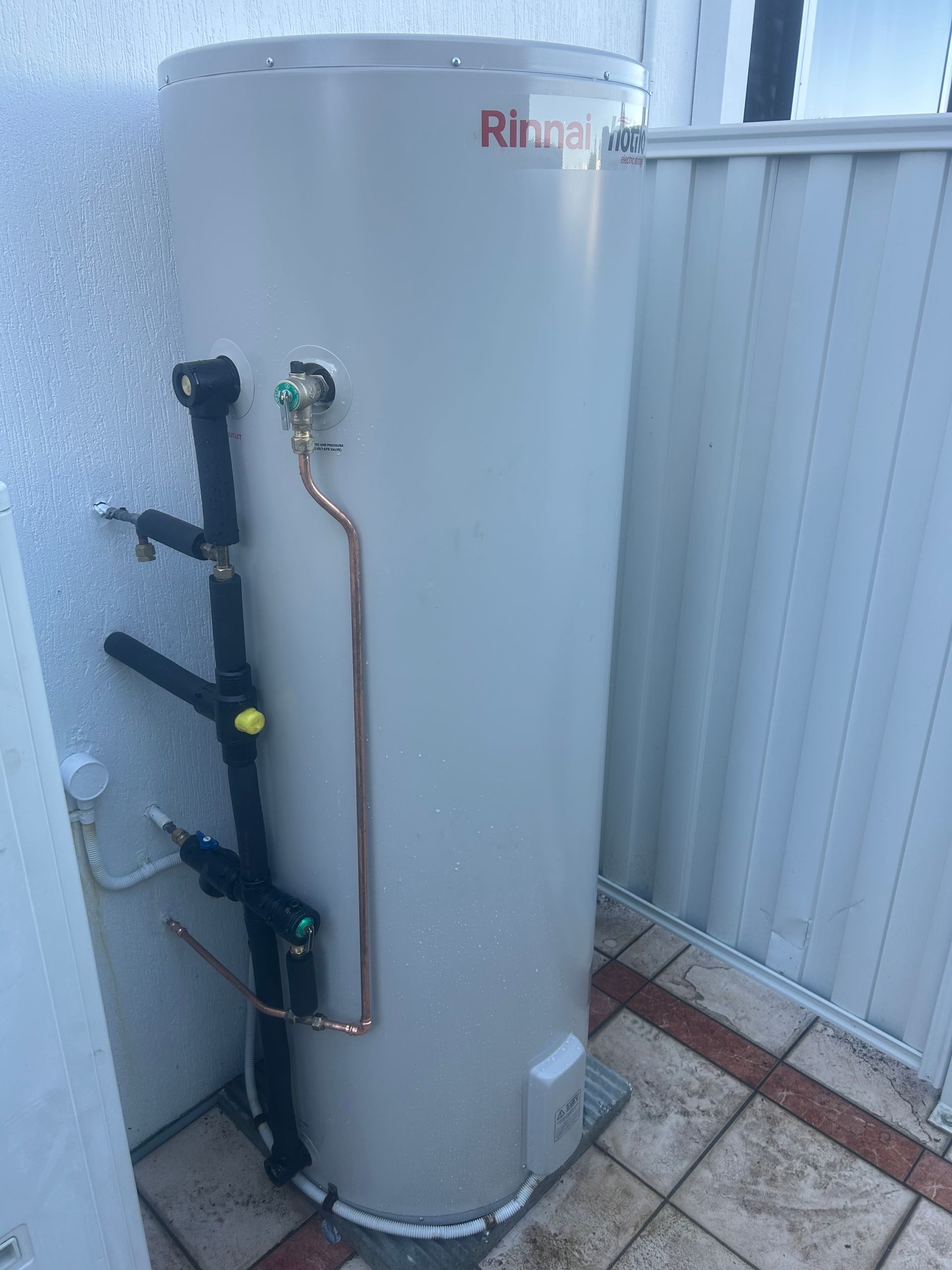 Water heater tank with exposed pipes and valves beside a corrugated metal fence on a tiled patio.
