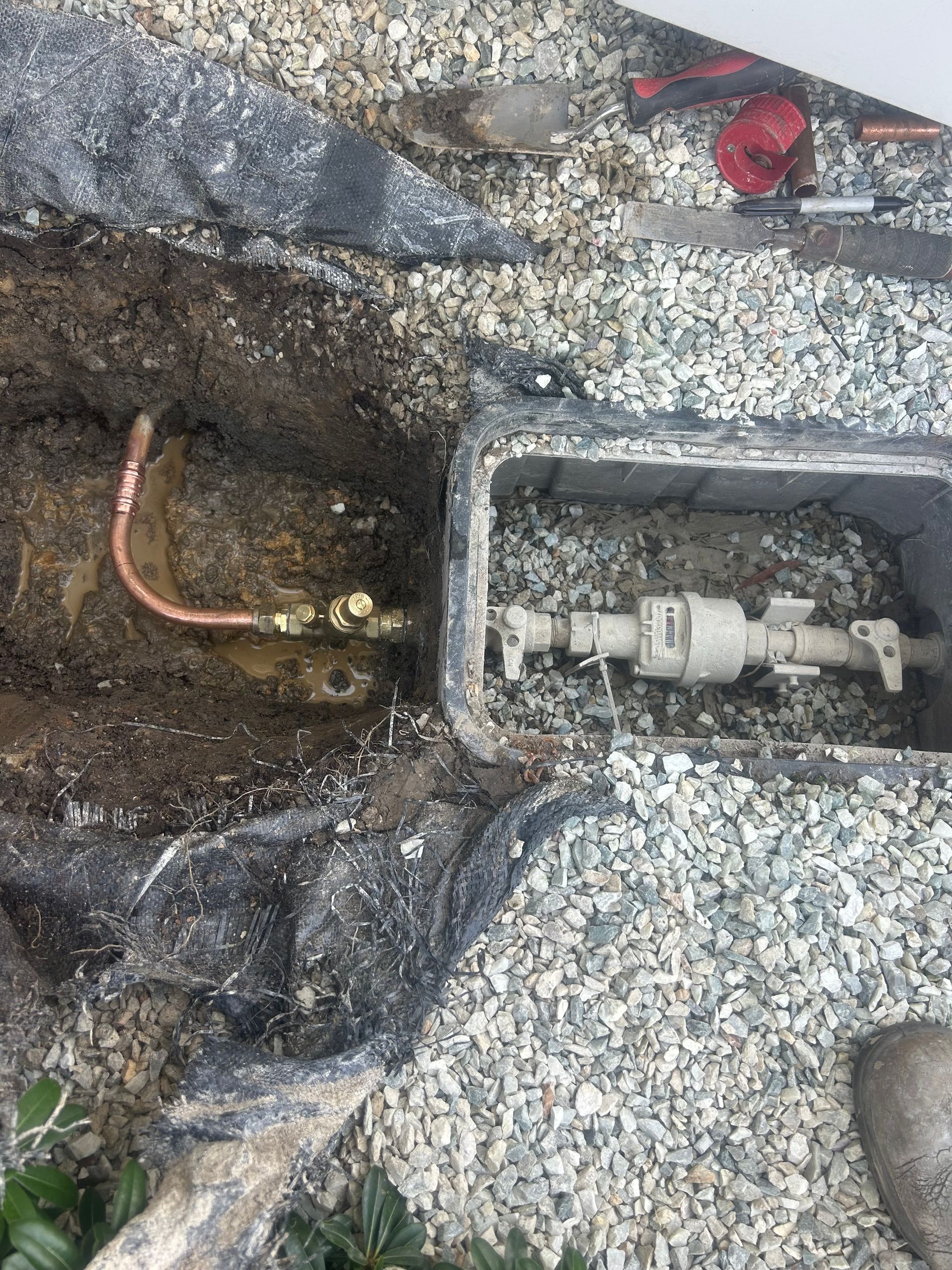Utility trench with exposed pipes and gravel around a concrete meter box. Includes a PRV.