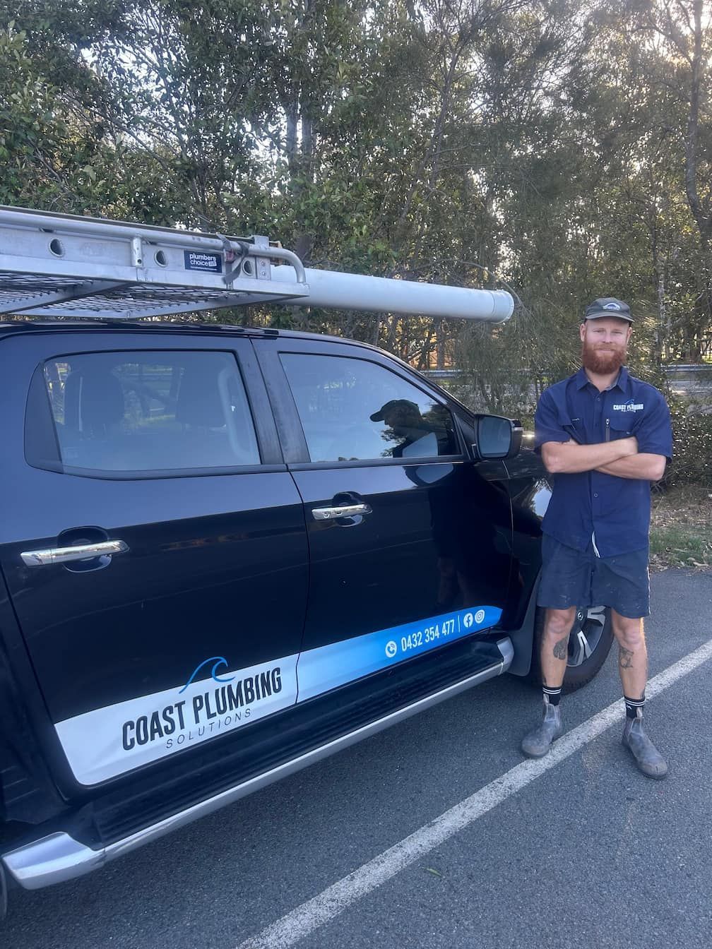 Plumber stands next to his work truck with ladder on top. The truck is parked. He has arms crossed.