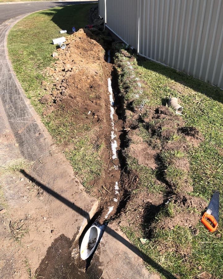 A trench dug for a white drainage pipe installation along a building's side on grass. A saw is present.