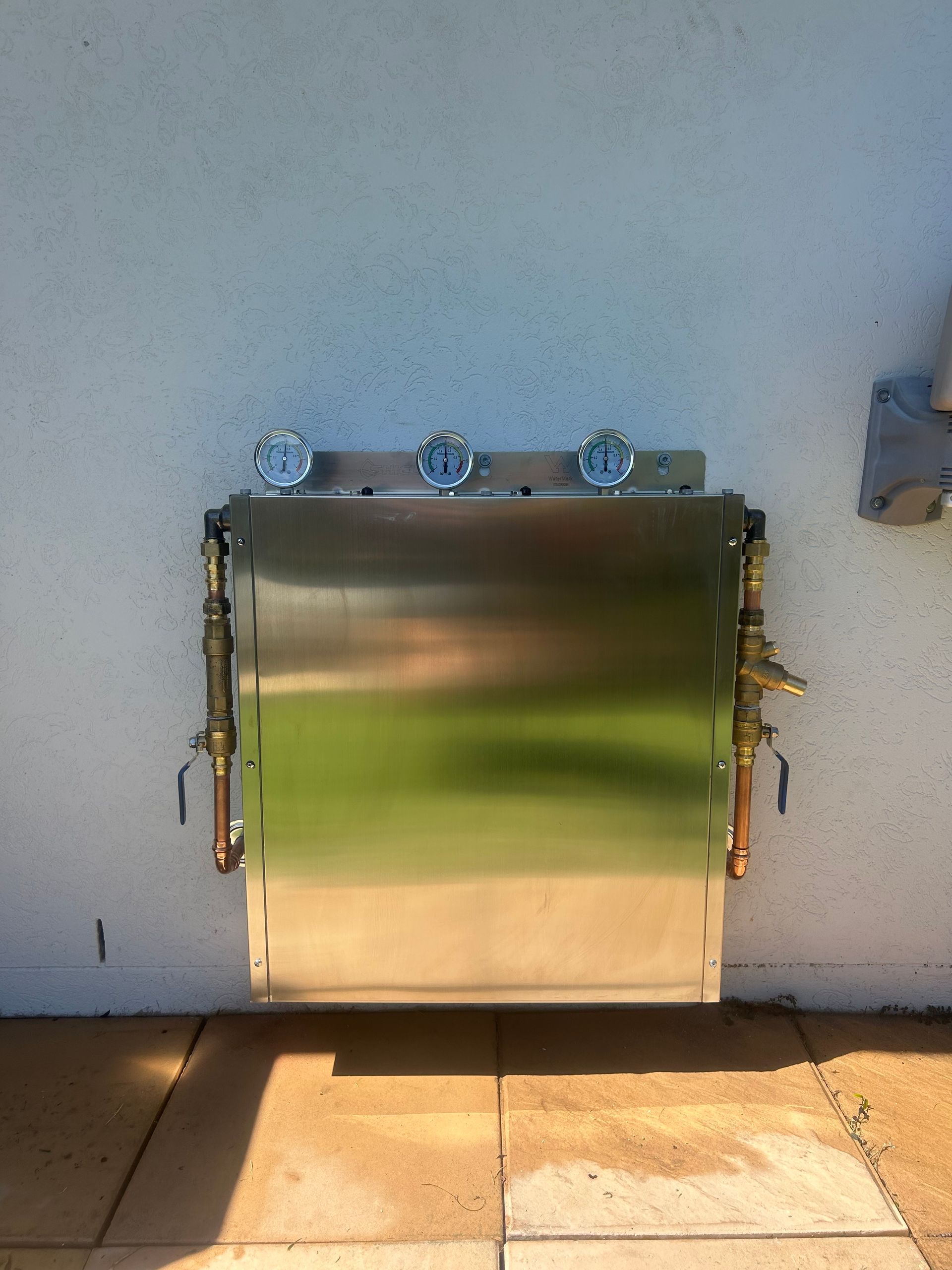 Stainless steel wall-mounted outdoor box with three valves against a stucco wall, above tiled ground.