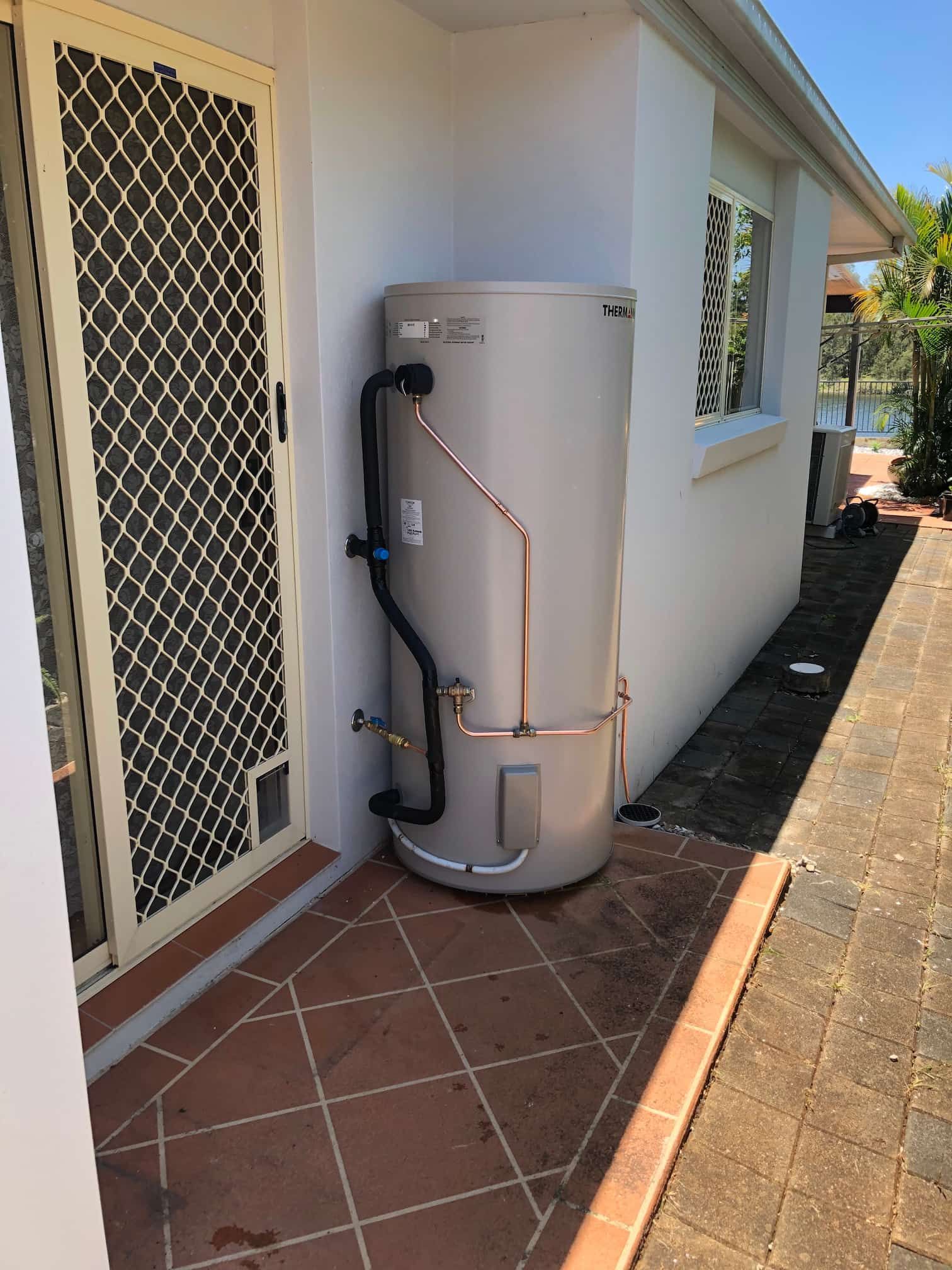 A cylindrical beige water heater installed outside on a concrete base, against a brick wall.