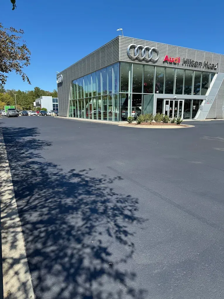 A modern glass-fronted Audi dealership building on a clear, sunny day, with a large, empty, freshly paved parking lot.