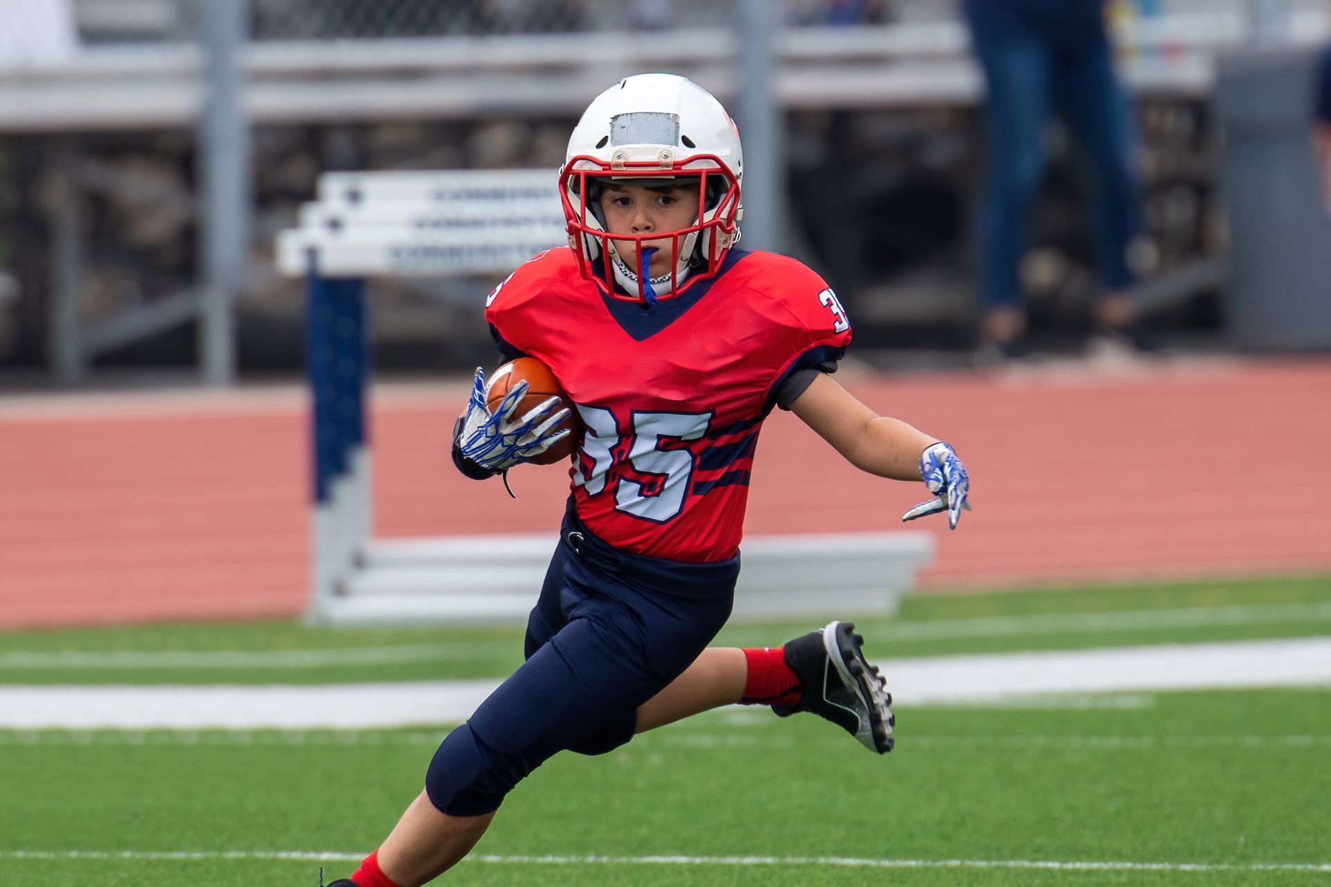 Youth football player runs with ball on a green field, wearing red and blue uniform.