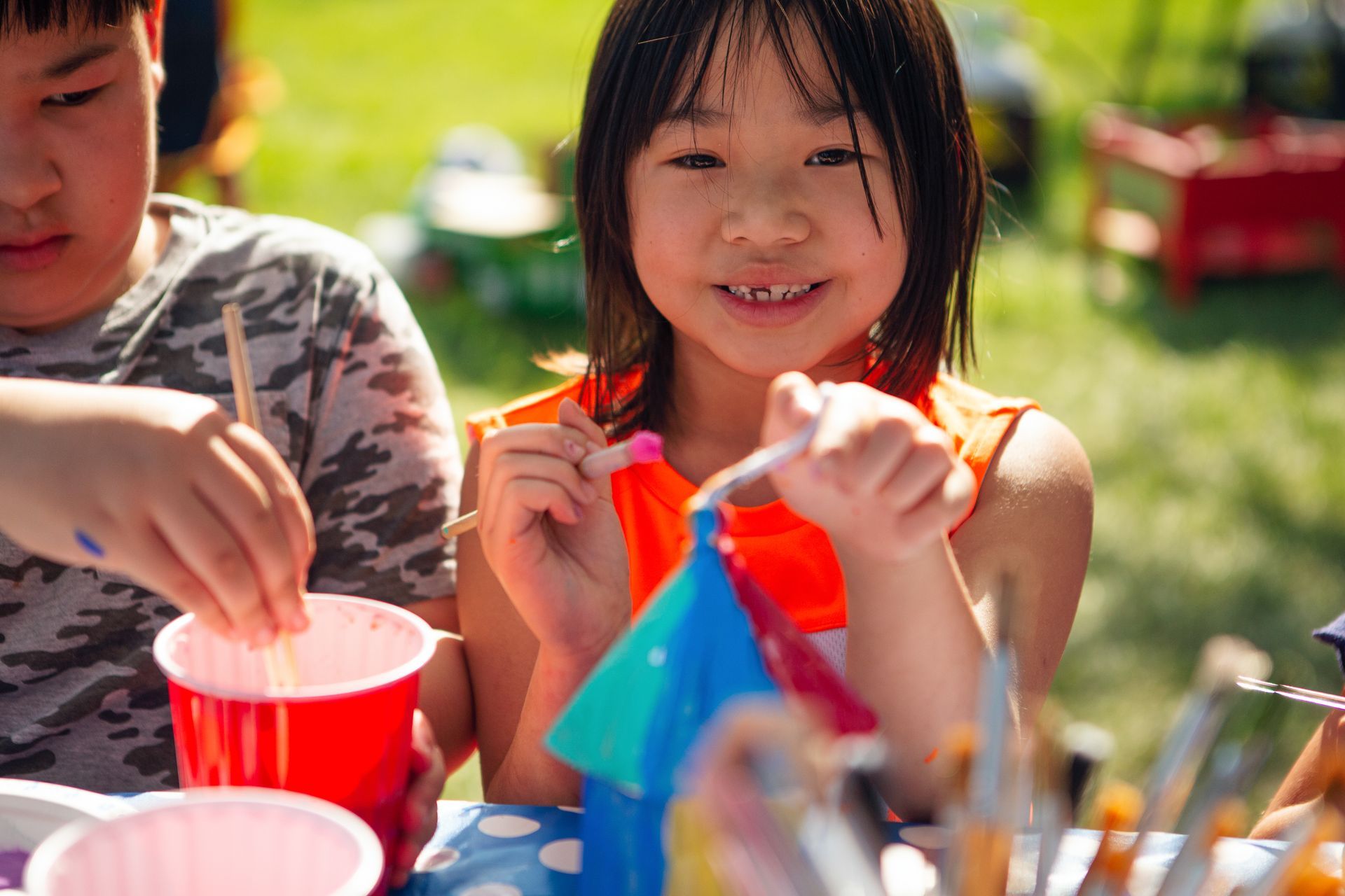 A young girl is sitting in a park working on a colorful art project. She is smiling.