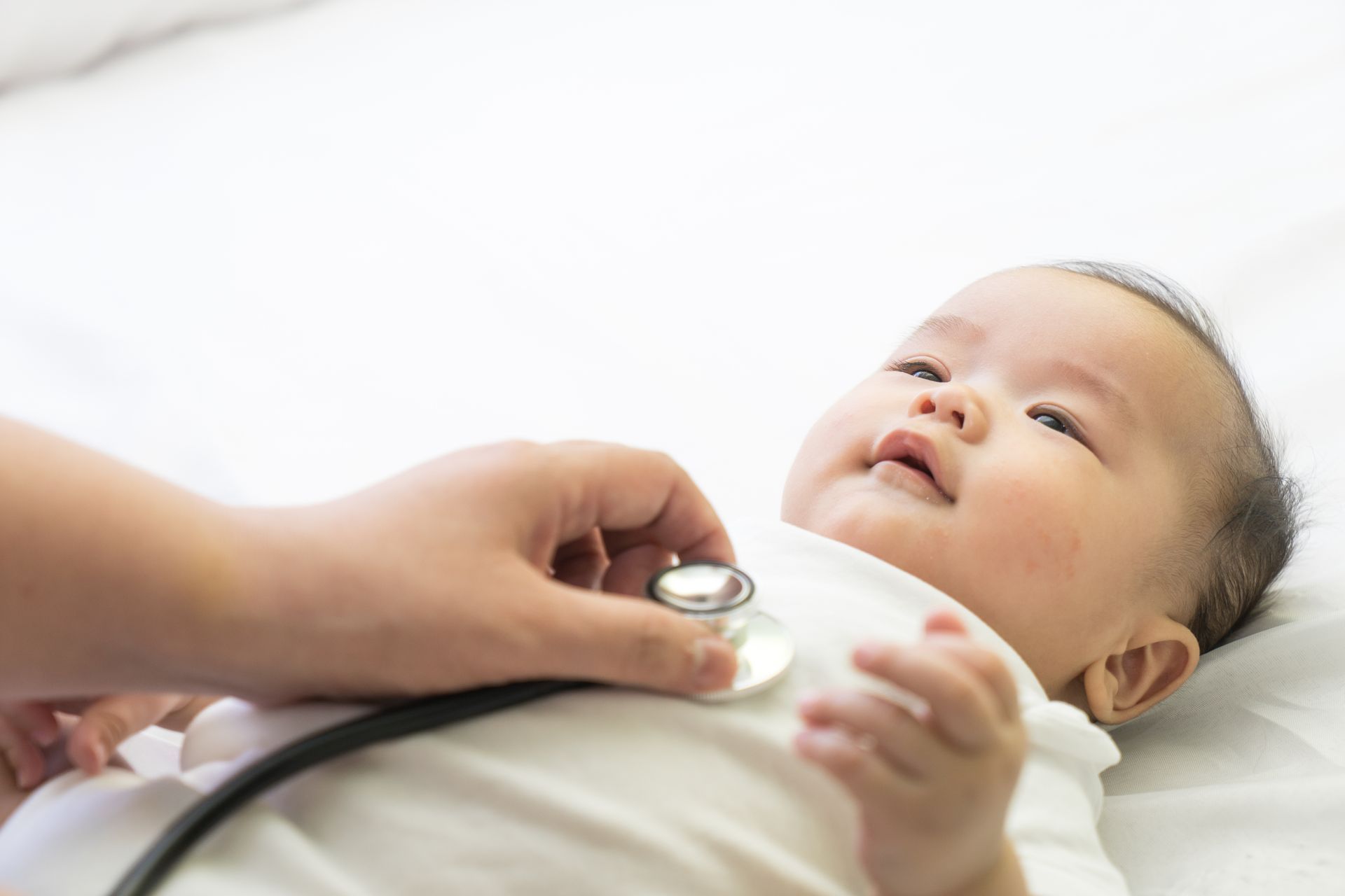 Doctor listening to baby's chest with a stethoscope; baby is on a white surface, looking upward.