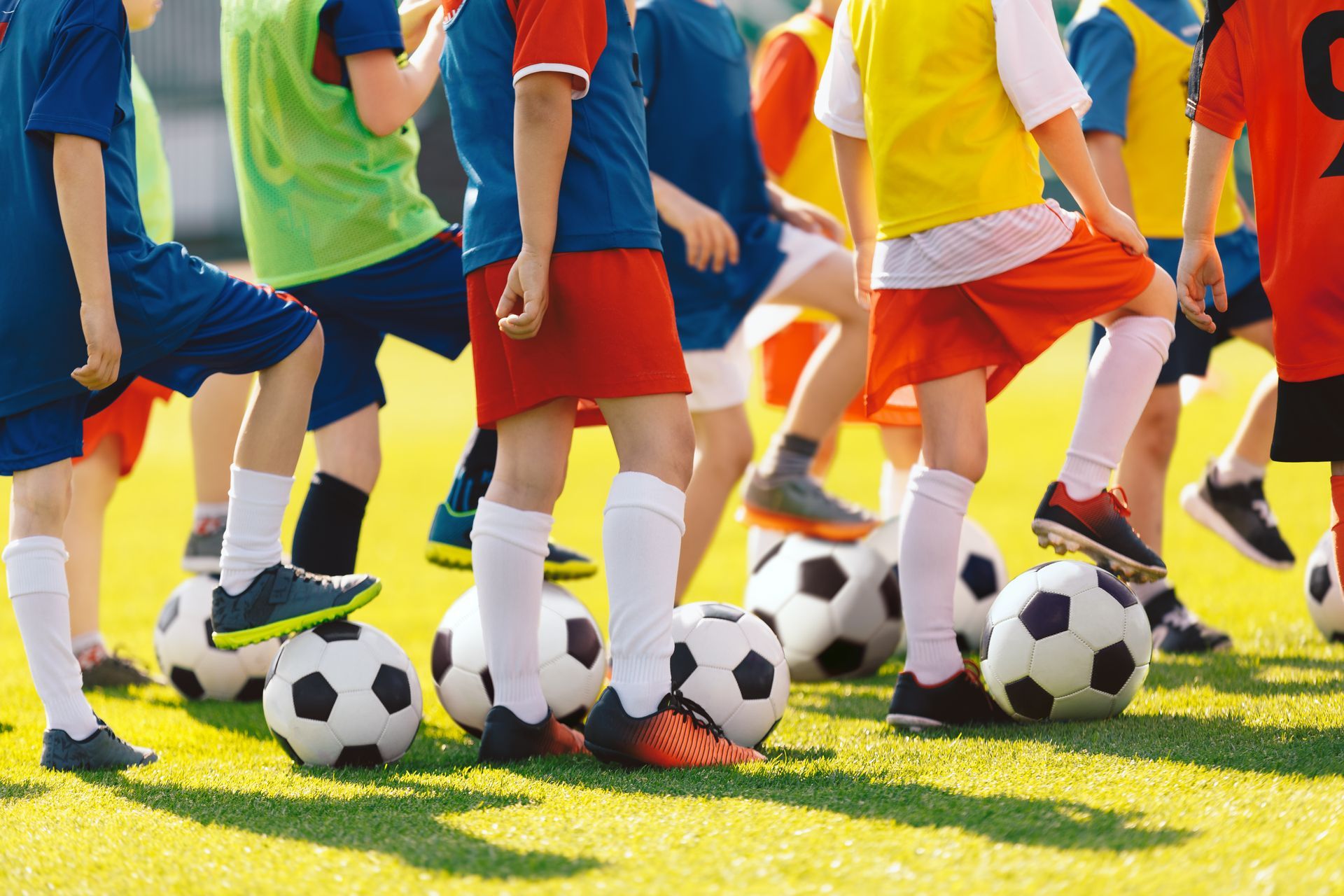 Children in soccer uniforms stand with feet on soccer balls on a green field.