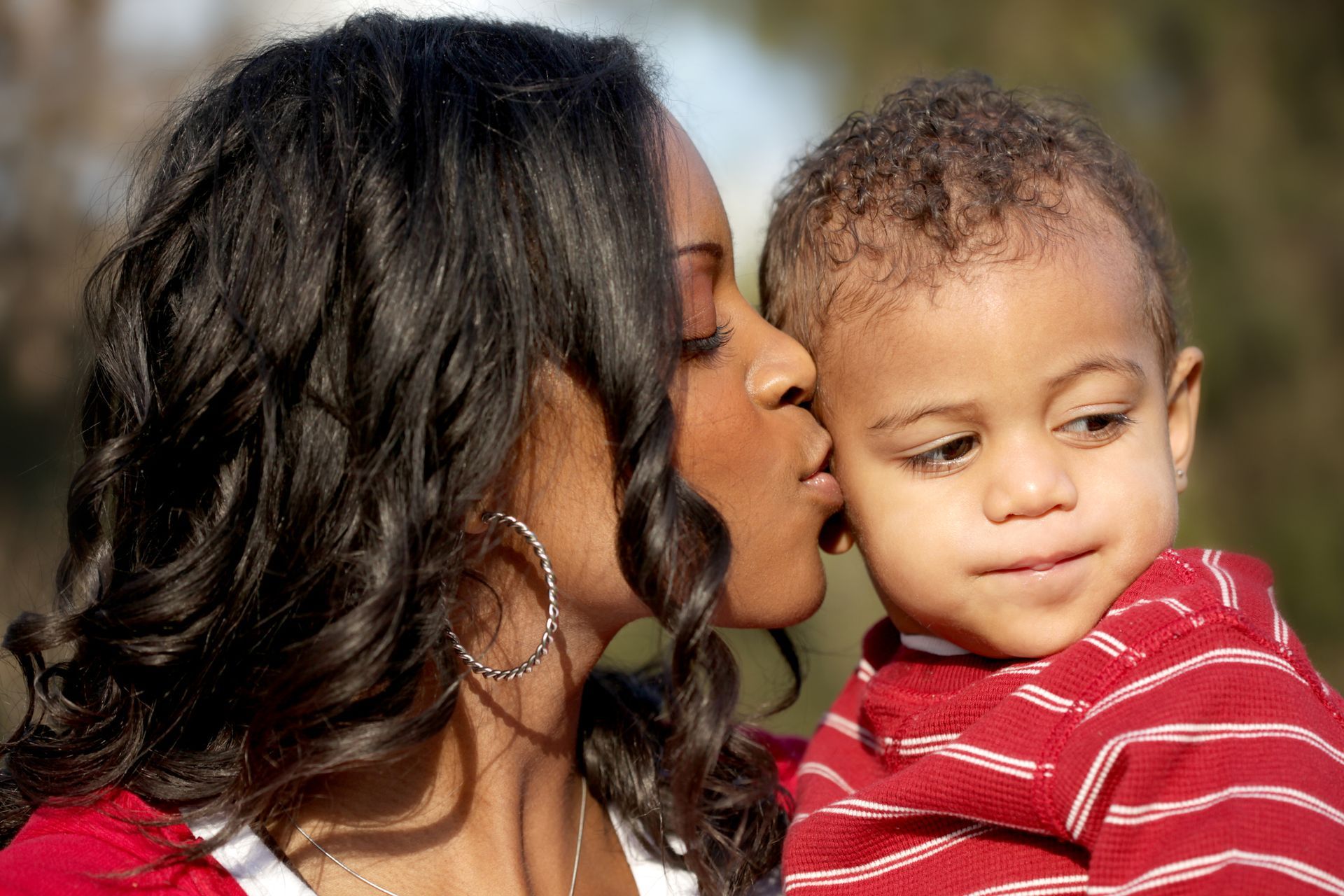 Mother holding her young child, kissing him on the cheek. They are in a park.