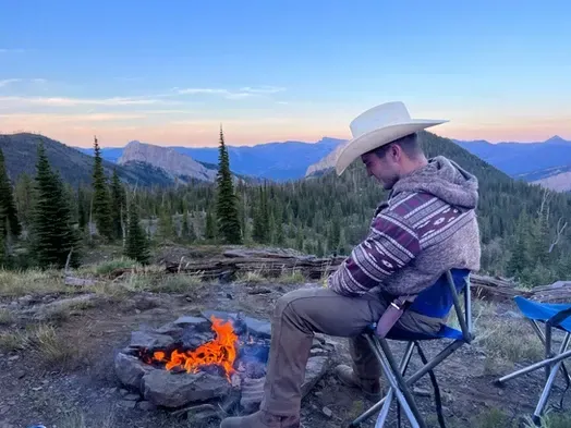 A man in a cowboy hat is sitting in front of a campfire.