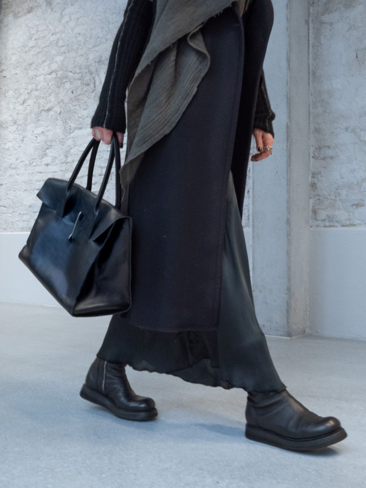 Eine Frau in schwarzem Kleid und Stiefeln trägt eine große schwarze Tasche und geht.